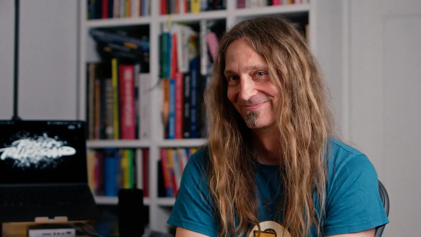 Marc Thiele smiling in front of a full bookshelf, a laptop with the beyond tellerrand lettering beside him