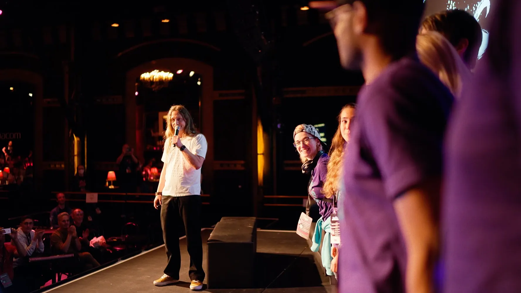 Marc Thiele speaking into a microphone on stage, crew members in purple shirts beside him and the audience filming on their phones