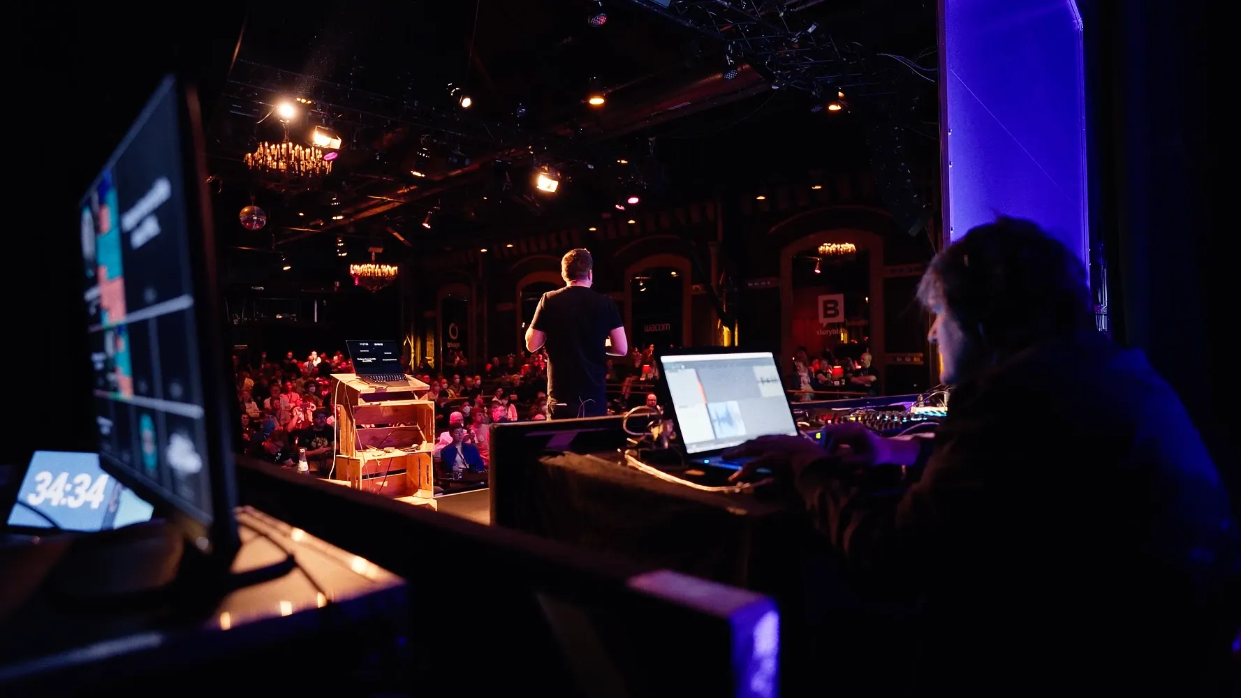 A technician at the laptop and mixer at the side of the hall, a monitor counting 34:34, while a speaker gives a talk on stage